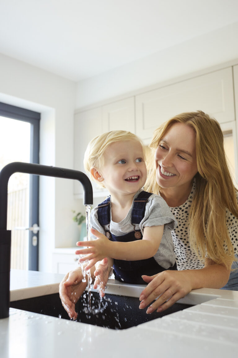 A woman and a young child are smiling while washing the child's hands under a running faucet in a bright kitchen. The woman has long hair and is wearing a polka-dot shirt, and the child is in a gray shirt with overalls.