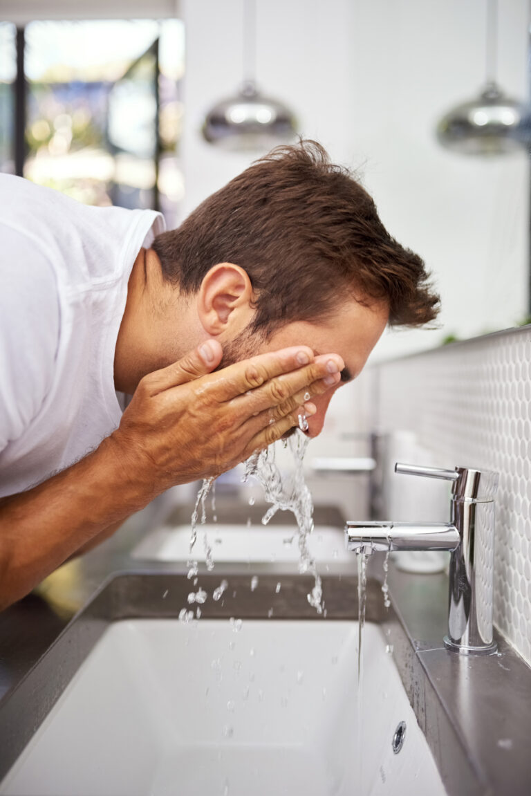 A man wearing a white shirt splashes water on his face over a modern sink. The bathroom features a white tiled wall and stainless steel fixtures. Two pendant lights can be seen in the blurred background.