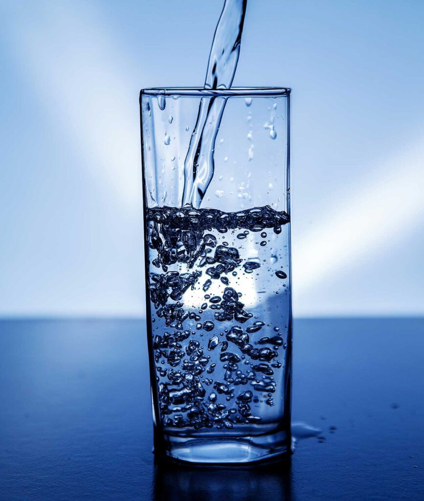A clear glass is being filled with water, creating bubbles as it pours. The background is a gradient of light and dark blue, highlighting the clarity of the water and the glass.